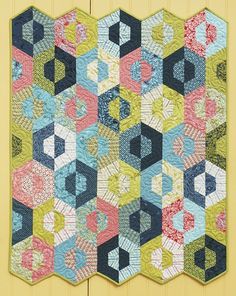 a quilted wall hanging on the side of a yellow wooden building with blue, green and pink hexagons