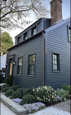 a gray house with black shutters on the front and windows, surrounded by flowers