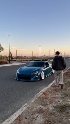 a man standing next to a blue sports car on the side of a road in front of a parking lot