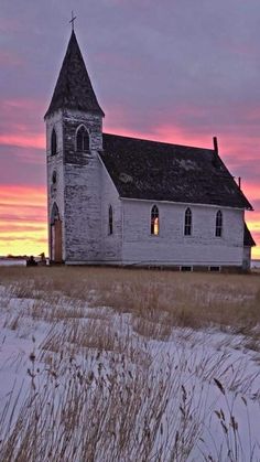 an old church sits in the middle of a snow covered field at sunset with tall grass