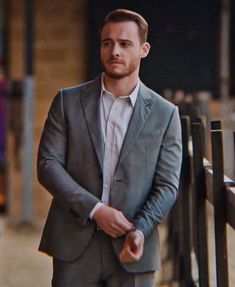 a man in a gray suit and white shirt standing next to a wooden fence with his hands in his pockets