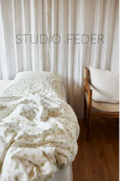 an unmade bed sitting next to a wooden chair in front of a white curtain