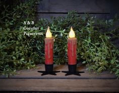 two red candles sitting next to each other on top of a wooden shelf with greenery in the background