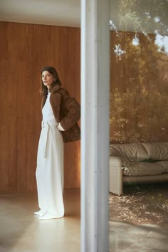 a woman in a long white dress and brown jacket is standing near a wall with wood paneling