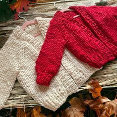 two knitted sweaters sitting on top of a wooden bench next to autumn leaves