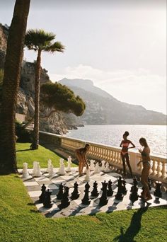 two women playing chess on the side of a road next to some water and palm trees