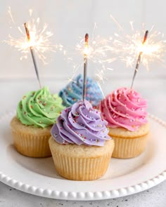 three cupcakes with colorful frosting and sparklers in the middle on a white plate