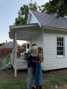 a man and woman standing in front of a small white house with a gray roof