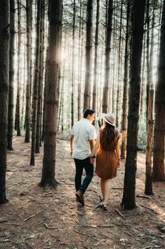 a man and woman walking through the woods in front of some tall trees, holding hands