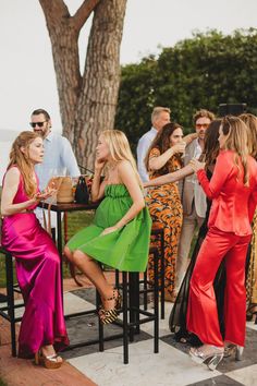 two women sitting at a table talking to each other in front of people standing around