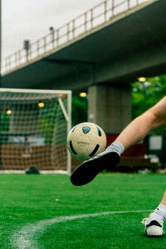 a man kicking a soccer ball on top of a lush green field with a bridge in the background