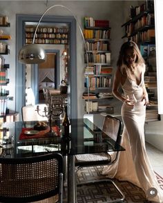 a woman is standing in front of a table with chairs and bookshelves behind her