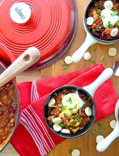 three bowls of chili and beans with spoons on a table next to two red casserole dishes