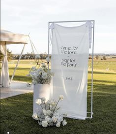 a white sign sitting on top of a lush green field next to a vase filled with flowers