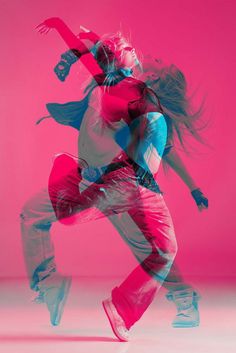 a woman in pink and blue is dancing on a pink background with her hair blowing in the wind