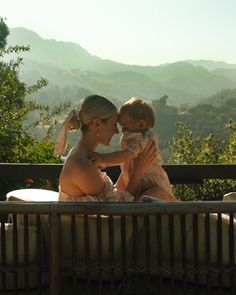 a woman holding a baby while sitting on top of a wooden bench in front of mountains