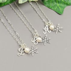 three silver necklaces with snowflakes and initial charms on them next to a green leaf