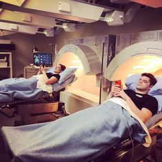 two men laying in hospital beds looking at their cell phones while one man takes a selfie with his phone