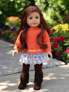 a doll with long brown hair wearing an orange sweater and white lace skirt is standing in front of flowers