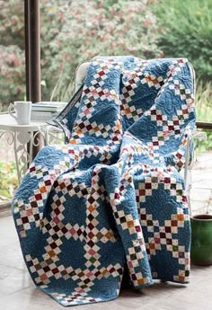 a chair with a quilt on it sitting in front of a window next to a potted plant