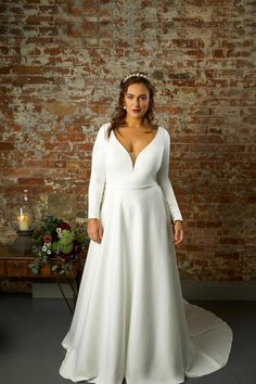 a woman standing in front of a brick wall wearing a white wedding dress with long sleeves