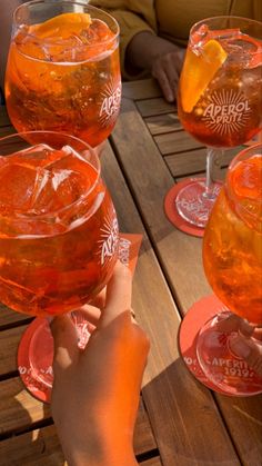 people holding up wine glasses with orange liquid in them on a wooden picnic table outdoors