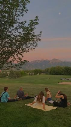 several people sitting on the grass in front of a tree at dusk, with mountains in the background