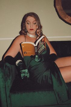 a woman in a green dress sitting on a couch reading a book with her legs crossed