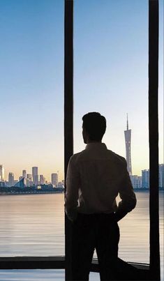 a man standing in front of a window looking out at the water and city skyline