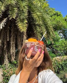 a woman holding up a drink in front of her face with palm trees behind her