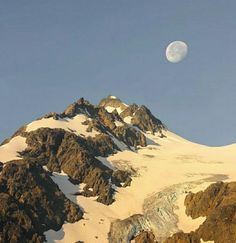 the moon is setting on top of a mountain with snow covered mountains in the foreground