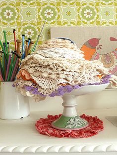 a white table topped with a cake covered in crocheted doily next to a cup filled with colored pencils