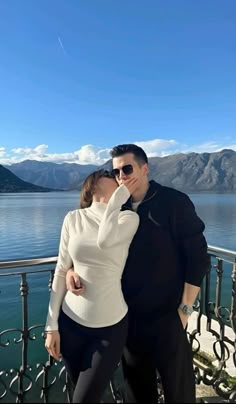 a man and woman standing next to each other on a balcony near the water with mountains in the background