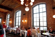 people sitting at tables in a restaurant with large windows and exposed brick walls, all looking out onto the street