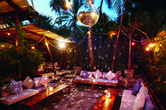 an outdoor seating area is lit up with lights and pillows on the floor, surrounded by palm trees