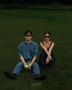 two people sitting on the grass in front of each other wearing sunglasses and holding flowers