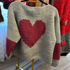 a sweater with a red heart on it is hanging in a store display rack next to other knitted clothing