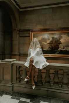 a woman in a white dress and veil sitting on a ledge next to a painting