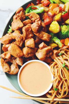 a plate full of chicken, noodles and vegetables with dipping sauce on the side next to some chopsticks