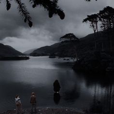 three people standing on the shore of a lake in front of some mountains under a cloudy sky