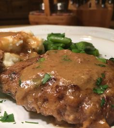 a white plate topped with meat covered in gravy next to potatoes and green beans