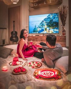 a man and woman sitting on a bed with pizzas, drinks and candles in front of the tv