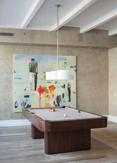 a pool table with a light hanging from it's side in front of a painting on the wall