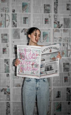 a woman standing in front of a wall covered in newspapers holding up a large newspaper