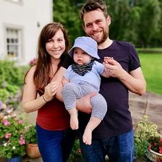 a man and woman holding a baby standing in front of a house with potted plants