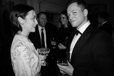 two people standing next to each other with wine glasses in their hands and one person wearing a tuxedo