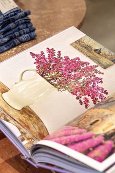 an open book with pink flowers on it sitting on a wooden table next to blue jeans