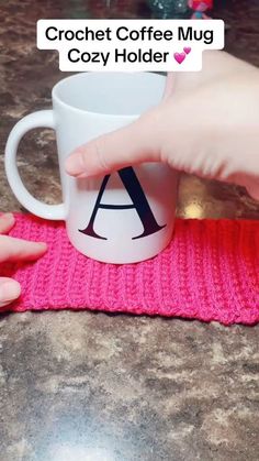 someone is holding a coffee mug in front of a pink knitted place mat with the letter a on it