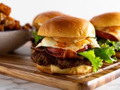 two burgers with bacon, egg and lettuce on a cutting board next to a bowl of tater tots