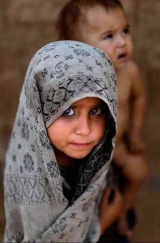 two young children with headscarves and scarves are looking at the camera while standing in front of a brick wall